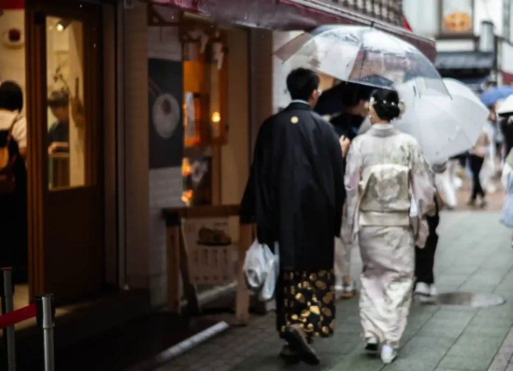 비 오는 아사쿠사 골목에서 투명 우산을 쓰고 걷는 기모노 입은 커플. 남성은 검은색 기모노, 여성은 흰색 기모노를 착용하고 있으며, 뒤로 상점의 따뜻한 조명이 보인다.