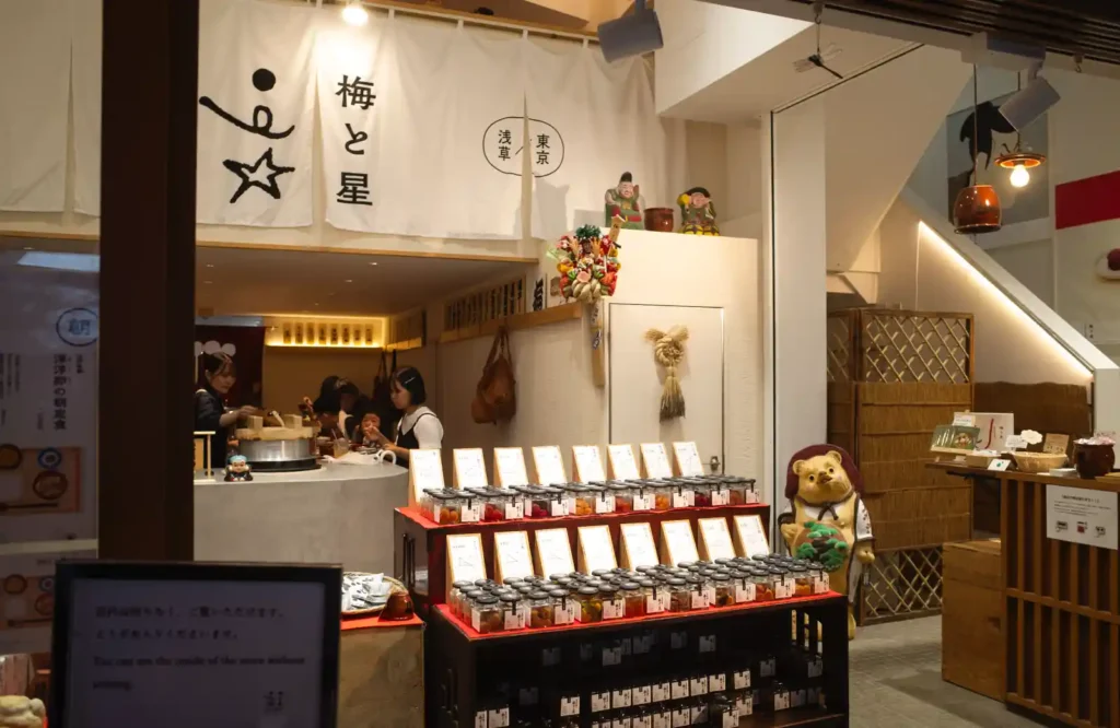 우메토호시 실내 전경. 입구에 '梅と星' 노렌이 걸려 있고, 중앙에는 다양한 종류의 우메보시가 담긴 병들이 빨간 진열대 위에 가지런히 놓여 있다. 왼쪽으로 직원들이 서빙하는 모습이 보이며, 벽면에는 전통 장식과 타누키 인형 등 아기자기한 소품들이 배치되어 있다.