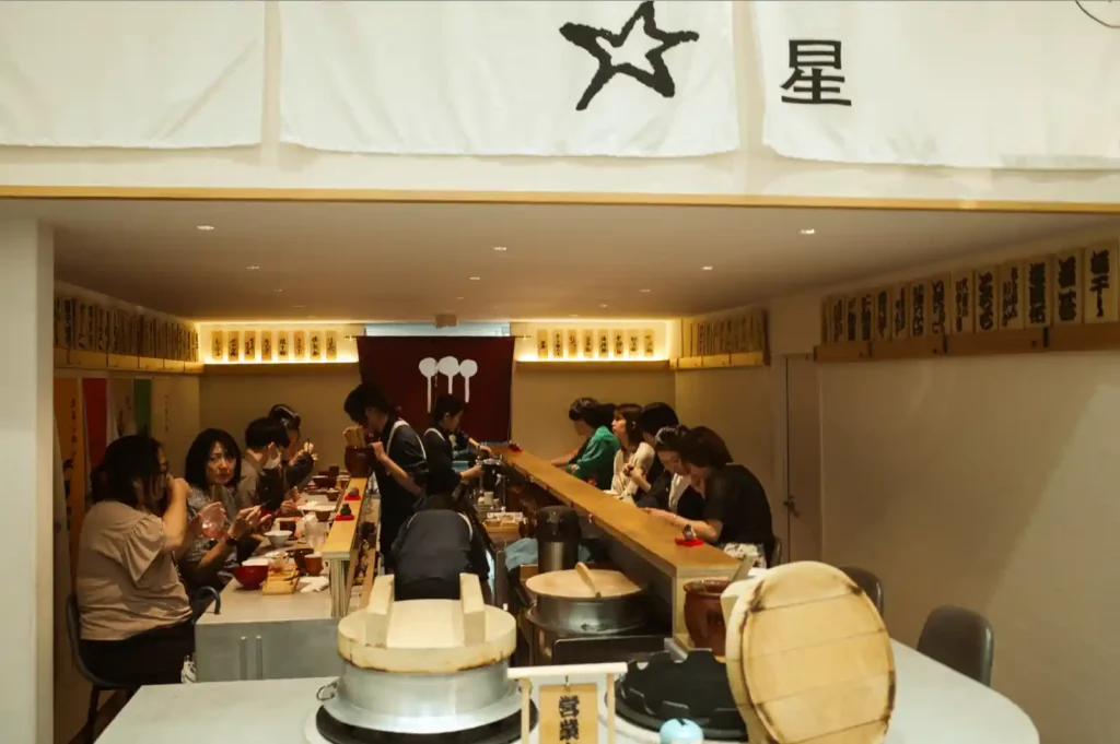 우메토호시 내부 좌석 배치. 중앙 통로를 기준으로 양쪽에 손님들이 앉아 식사하고 있으며, 직원들이 중앙에서 서빙하고 있다. 전경에는 흰색 하가마(전통 가마솥) 두 개가 보이고, 벽면에는 우메보시 종류가 적힌 메뉴판들이 걸려 있다. 천장의 '星'(별) 글자가 새겨진 노렌이 보인다.