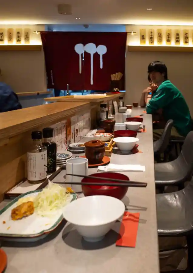 우메토호시 식사 중 카운터 풍경. 중앙 통로를 기준으로 양쪽에 손님들이 앉아 조용히 식사하고 있으며, 카운터 위에는 밥그릇, 된장국, 반찬 접시들이 정갈하게 놓여 있다. 뒤편 벽면에는 주걱 세 개가 그려진 빨간 노렌과 우메보시 종류가 적힌 메뉴판들이 보인다.