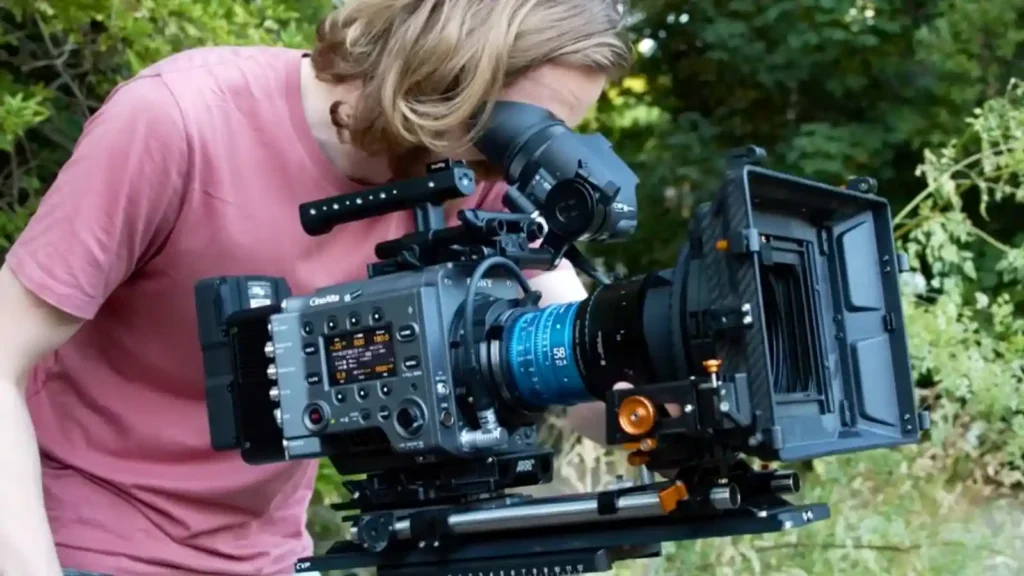 야외에서 전문 시네마 카메라 리그를 조작하는 영상 제작자. 시네마 카메라 바디에 58mm 시네 렌즈, 매트박스, 팔로우 포커스, 외부 모니터가 장착되어 있으며, 카메라는 레일 시스템 위에 마운트되어 있습니다. 촬영자는 뷰파인더를 통해 구도를 확인하고 있습니다.