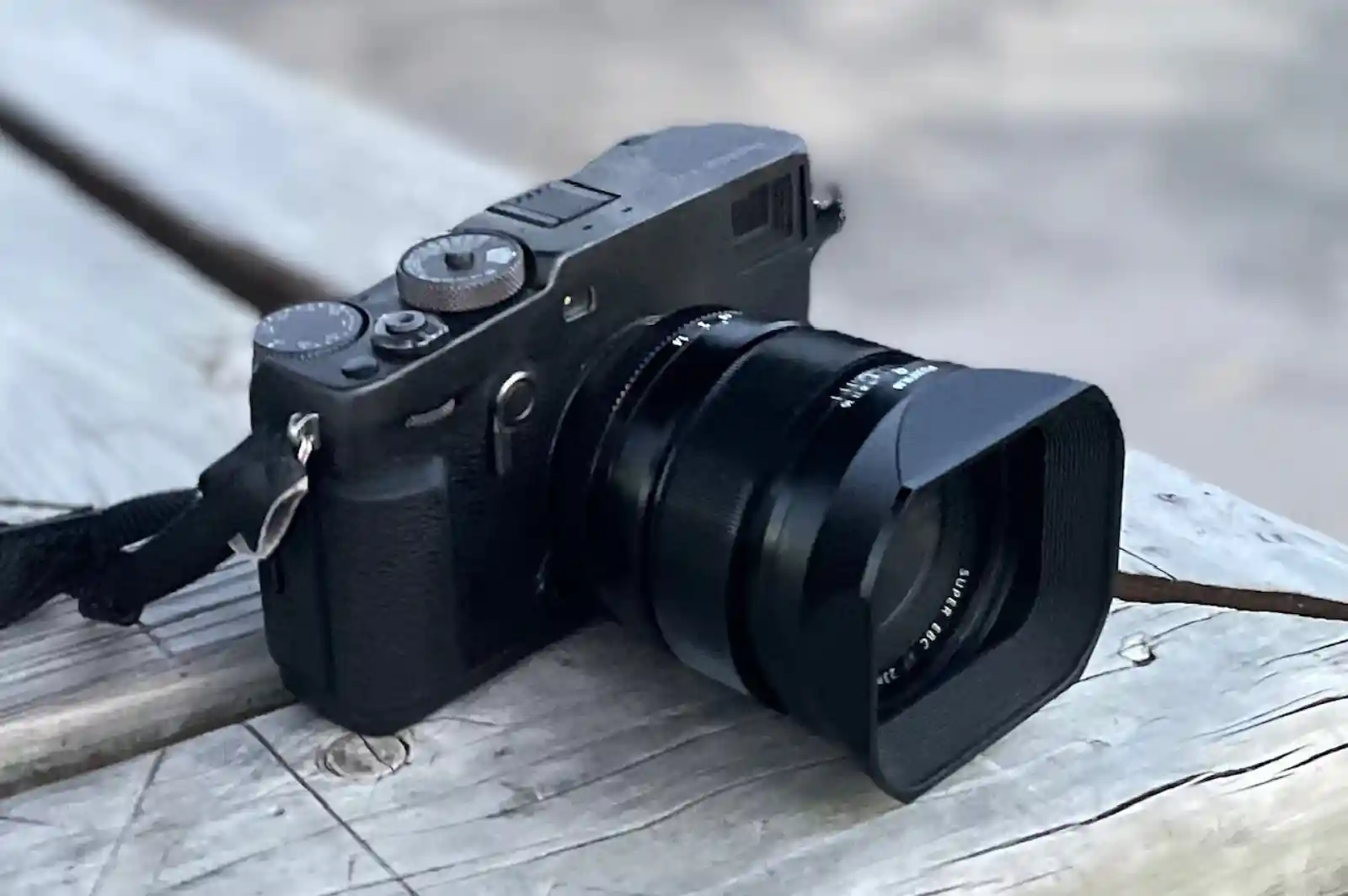 나무 위에 놓인 Fujifilm X-Pro3 블랙 바디. 스트랩이 걸려 있으며 후드가 장착된 XF 렌즈가 부착되어 있습니다.