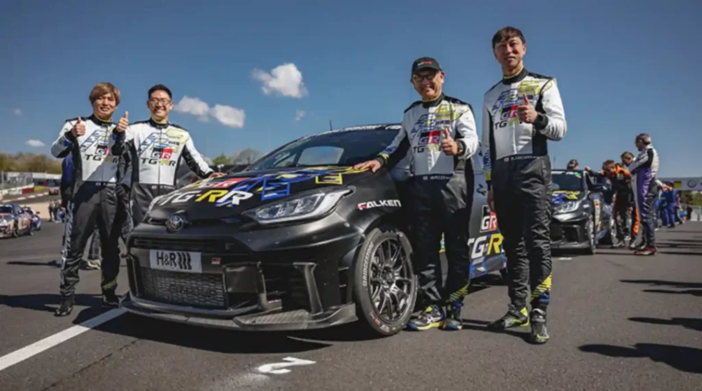 서킷에서 GR 야리스 레이싱카 앞에 레이싱 슈트를 입고 젊은 드라이버들과 엄지를 치켜들며 환하게 웃는 도요다 아키오 모리조