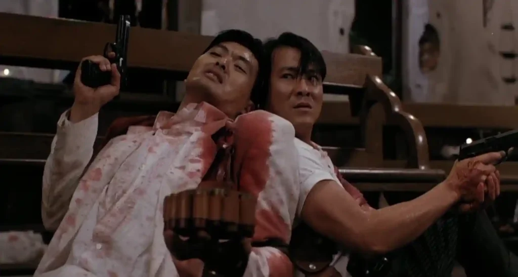 영화 첩혈쌍웅의 한 장면, 피로 얼룩진 흰 셔츠를 입은 주윤발이 권총을 들고 있고 이수현이 그를 뒤에서 지탱하고 있다