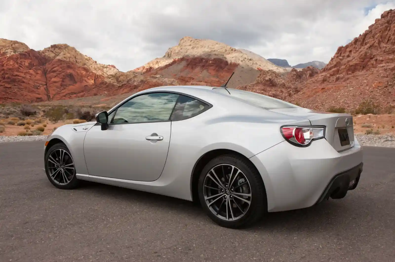 사막 도로에 주차된 실버 색상의 Scion FR-S 스포츠카 측후면. 붉은 암석 산맥을 배경으로 2도어 쿠페의 날렵한 실루엣과 스포츠 휠이 돋보인다