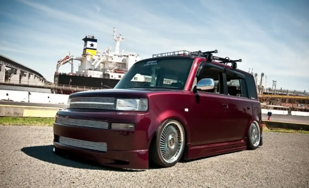 항구를 배경으로 선 버건디 색상의 커스터마이징된 Scion xB. 로우다운 서스펜션, 크롬 와이어 휠, 루프랙 등 개성 있는 튜닝이 적용되어 싸이언의 커스터마이징 철학을 보여준다