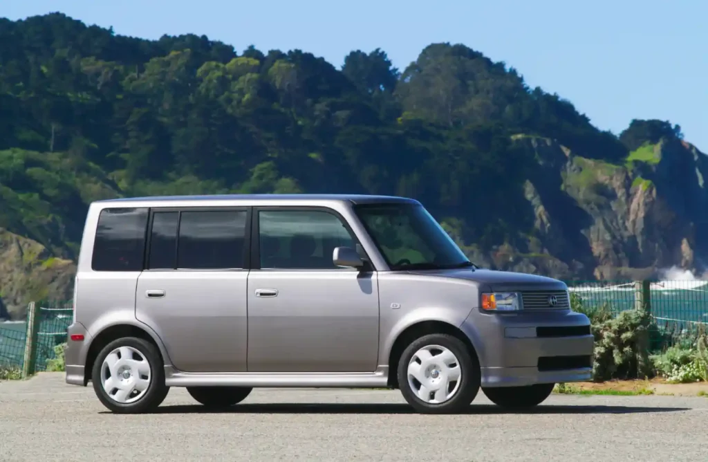 바다가 보이는 해안도로에 주차된 실버 색상의 1세대 Scion xB. 특징적인 박스형 디자인과 흰색 휠캡이 보이는 순정 상태의 5도어 해치백