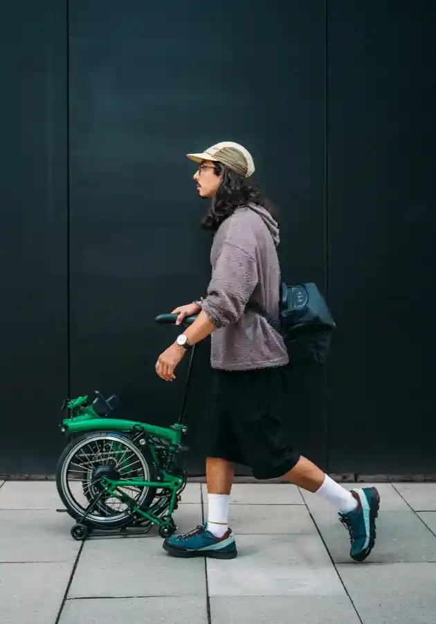 접힌 브롬톤 전기자전거를 핸들바로 끌며 보도를 걷고 있는 남성 — SuperRoll 이동 방식 실사용 장면