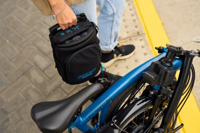 브롬톤 전기자전거 배터리 팩을 손으로 분리하는 장면, 접힌 상태의 블루 컬러 브롬톤 일렉트릭과 함께