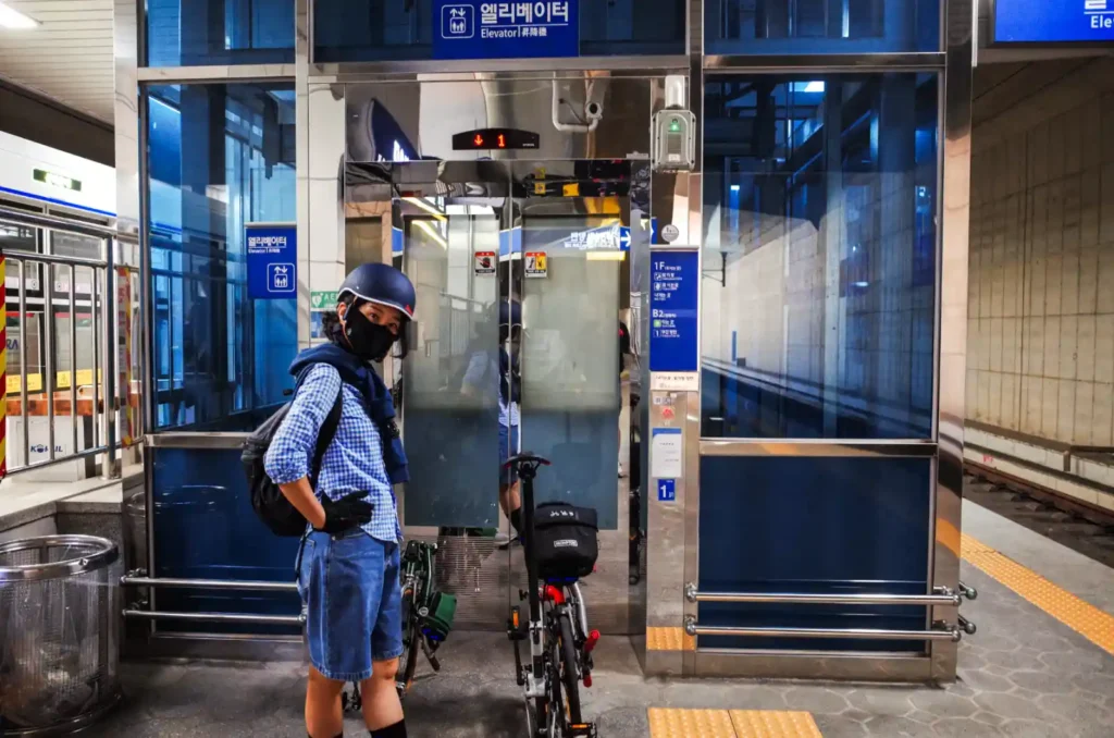 지하철역 엘리베이터 앞에서 브롬톤 자전거를 끌고 서 있는 여성. 어반 헬멧을 착용하고 있다.