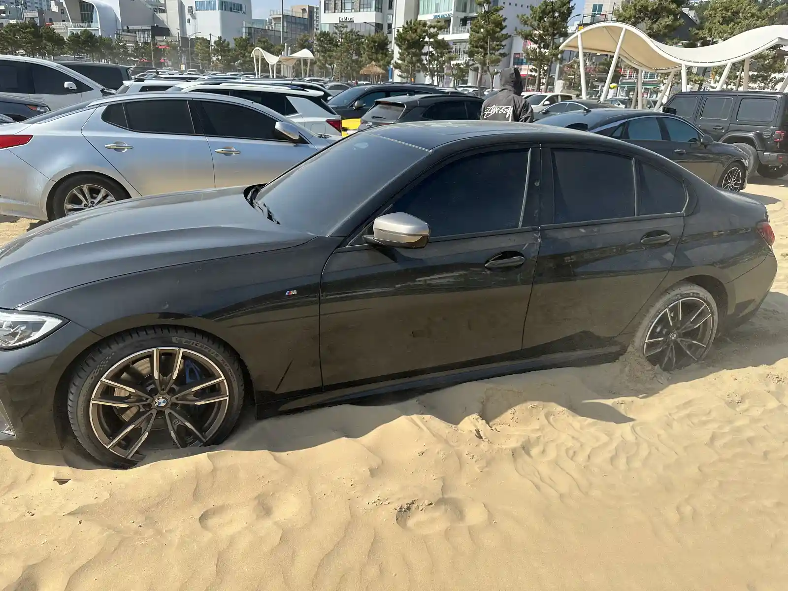 포항 영일대해수욕장 공용주차장 모래 구간에 빠진 BMW M340i 측면 전경 — 뒷바퀴가 모래 속에 매몰된 상태, 주변 일반 차량들과 대비
