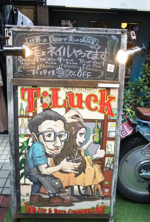 시모키타자와 미용실 T:Luck 입간판. 상단 칠판에 맥주와 네일 서비스 안내가 적혀 있고, 하단에 'HAIR CUT & BEER COMMUNICATION' 문구와 일러스트가 그려져 있다.