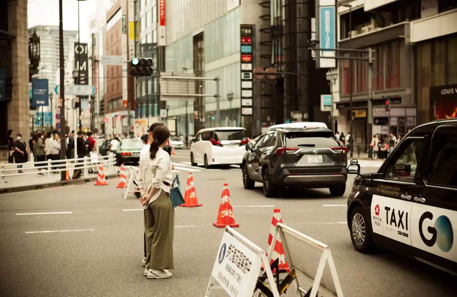 도쿄 신주쿠 도심 거리에서 보행자가 횡단보도 앞에 서 있고, 택시와 차량이 지나가는 붉은 보도블록의 오후 풍경