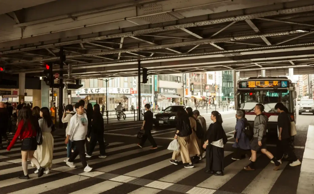 메구로역 앞 고가 아래 횡단보도에서 사람들이 건너는 모습, 오른쪽에 目黒駅 행 버스가 정차해 있고 뒤로 吉そば 간판이 보이는 오후 풍경