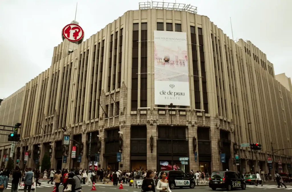 신주쿠 이세탄 백화점 본관 외관, 아르데코 양식의 베이지색 건물 외벽에 Clé de Peau Beauté 대형 광고가 걸려 있고 앞 교차로에 사람들이 붐비는 모습