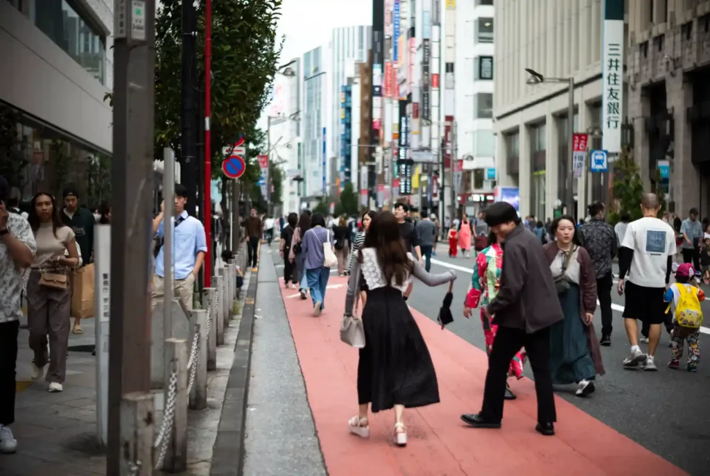 신주쿠 도심 붉은 보도블록 위로 다양한 사람들이 걷고 있는 오후 거리 풍경, 뒤로 간판들과 삼井住友銀行 건물이 보이는 모습