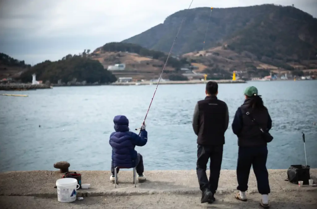 욕지도 방파제에서 낚시 중인 세 사람의 뒷모습, 배경으로 욕지항과 산 능선이 펼쳐지는 겨울 풍경