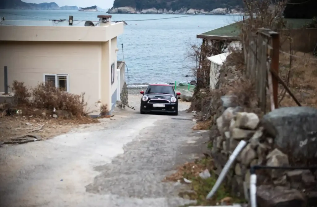 욕지도 어촌 마을 좁은 골목길을 내려오는 검정 미니쿠퍼, 끝으로 한려수도 바다와 기암괴석이 보이는 풍경