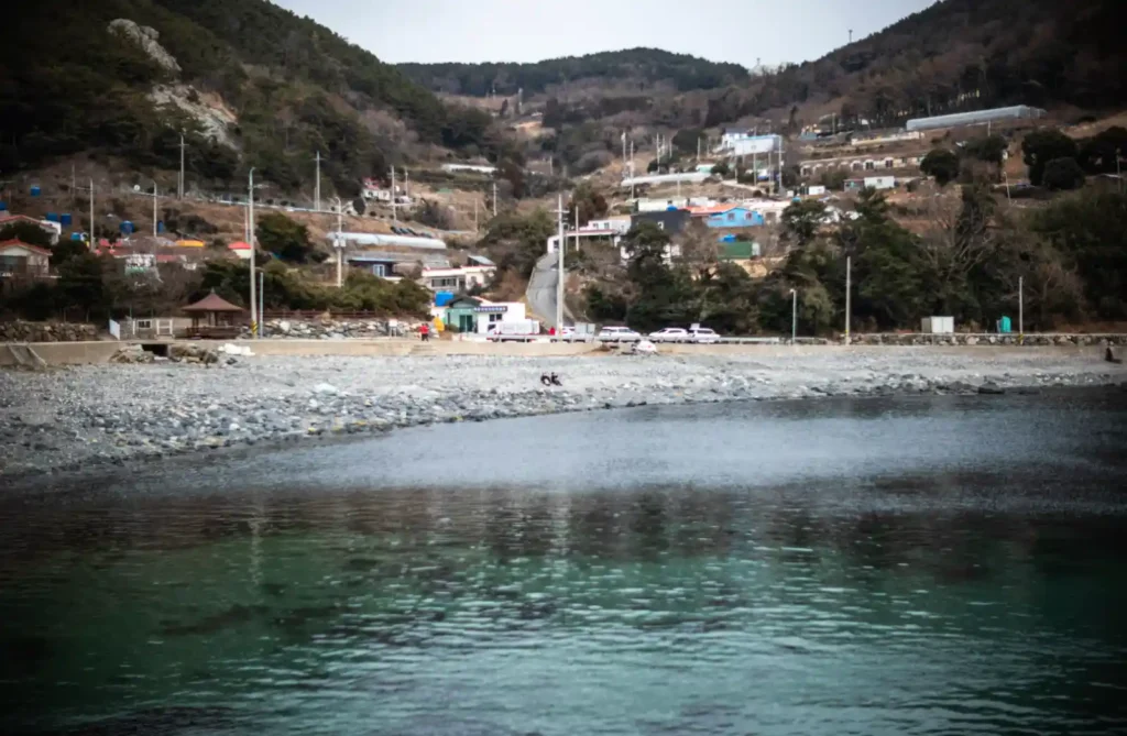 욕지도 덕동해수욕장 전경, 몽돌 해변과 맑은 바닷물 너머로 가파른 비탈에 자리 잡은 어촌 마을과 해안도로