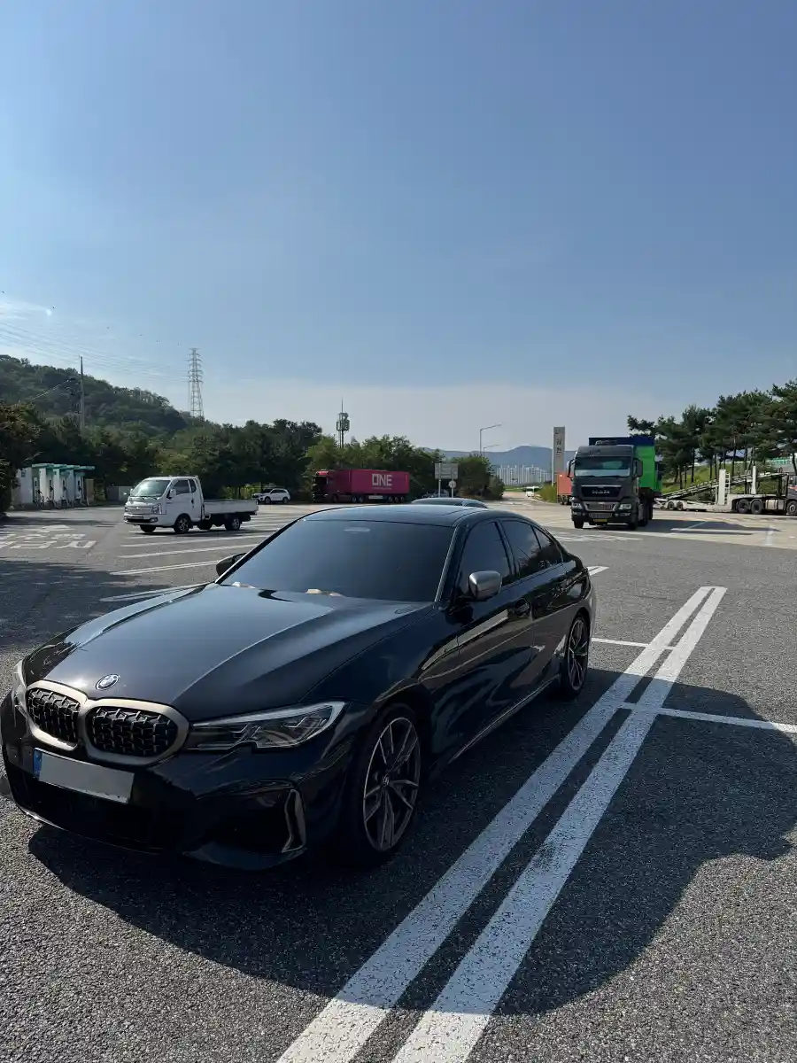 고속도로 휴게소에 세워진 검은색 BMW M340i 정면. 맑은 하늘 아래 대형 트럭들이 배경에 보인다.
