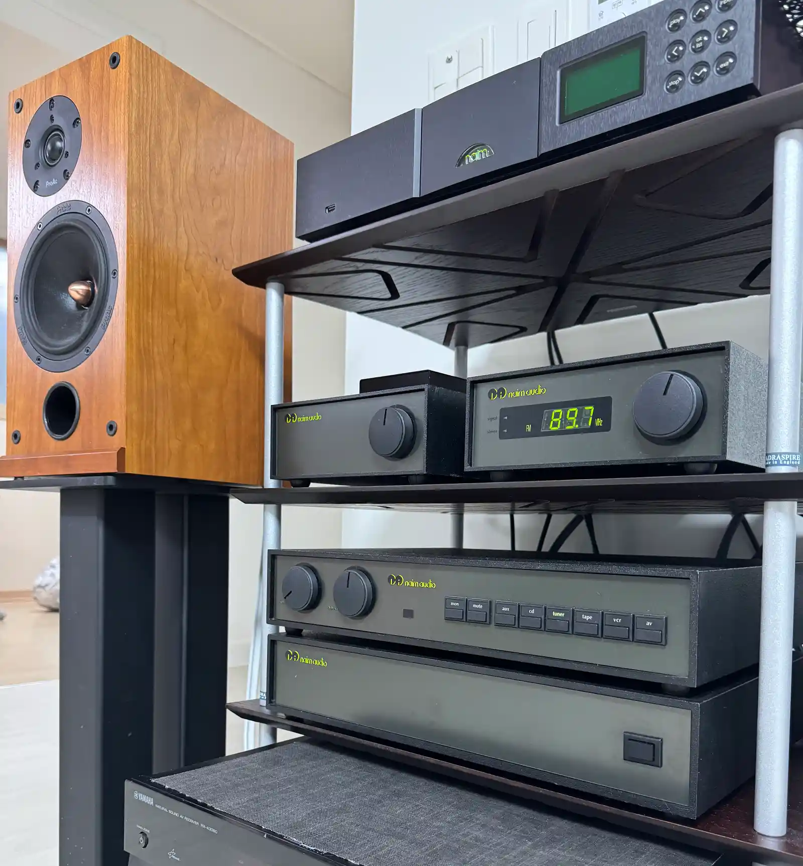 Naim 랙에 적층된 NDX, nDAC, NAT 01, NAC 102, NAP 180과 왼쪽 스탠드 위의 ProAc D2 클로즈업. Naim Audio 로고와 NAT 01의 89.7MHz 디스플레이가 선명하게 보입니다.