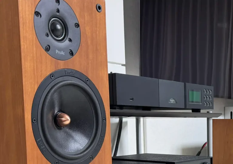 거실 랙 위에 놓인 Naim NDX 네트워크 플레이어와 왼쪽에 배치된 ProAc D2 체리 스피커. NDX 전면 디스플레이에 녹색 불이 켜져 있고, ProAc 로고와 트위터·우퍼 유닛이 선명하게 보입니다.