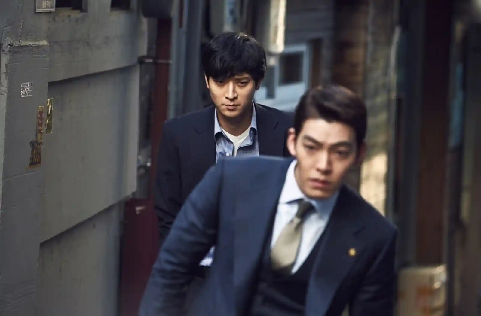마스터(2016) 스틸컷, 좁은 골목에서 정장 차림의 두 남성이 긴장된 표정으로 걷고 있는 장면, 앞사람은 넥타이를 매고 뒷사람은 날카로운 눈빛으로 따라가는 모습