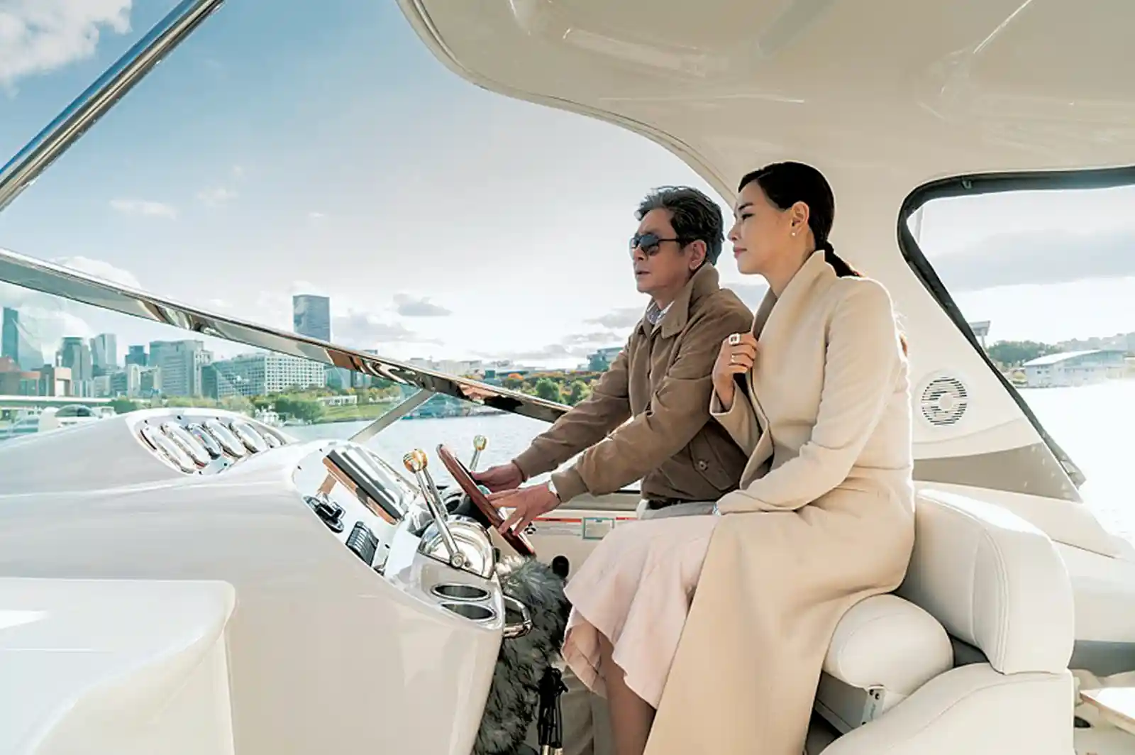 침묵(2017) 스틸컷, 선글라스를 쓴 중년 남성과 베이지색 코트 차림의 여성이 고급 요트 조종석에 나란히 앉아 정면을 바라보는 장면, 배경은 도심 스카이라인과 강