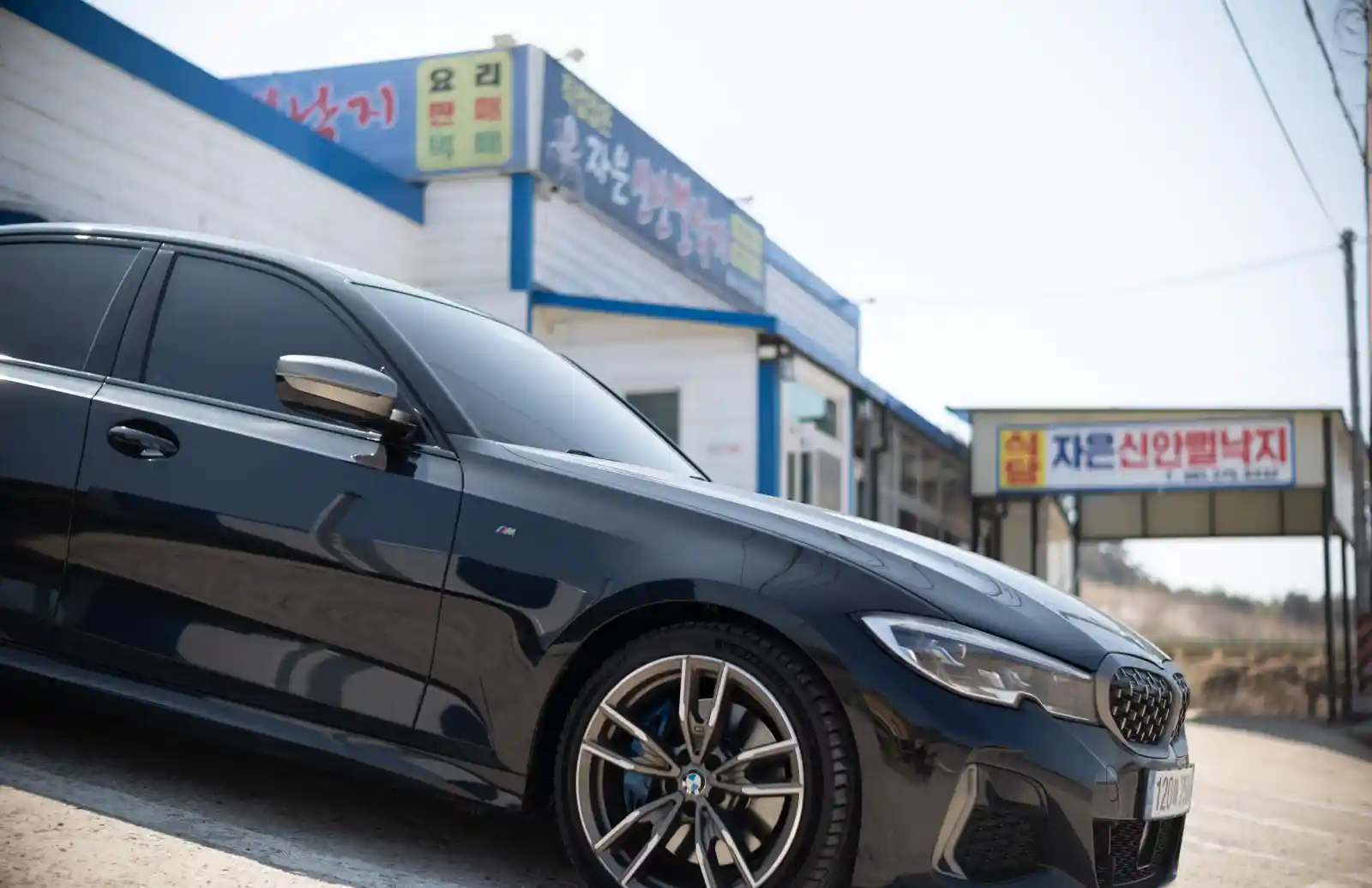 자은신안뻘낙지 식당 앞에 주차된 검정 BMW M340i 측면, 파란색 건물 외벽과 간판이 배경에 보이는 모습