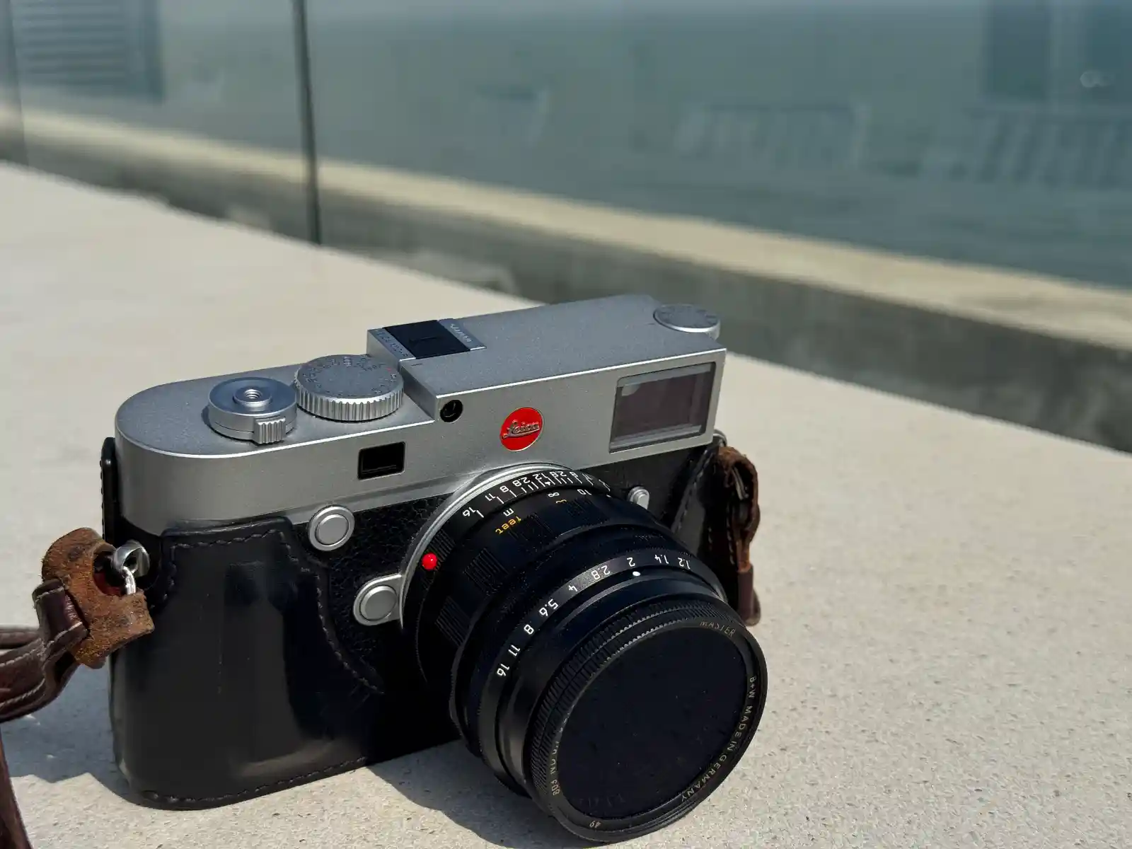 가죽 하프케이스와 브라운 스트랩이 장착된 Leica M10-R 실버 크롬 바디. 야외 콘크리트 바닥 위에 놓여 있으며 50mm 렌즈가 부착되어 있습니다.