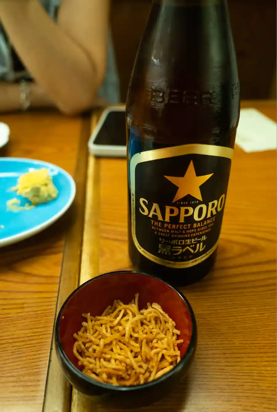 사라시나 호리이 테이블 위 삿뽀로 黒ラベル 맥주병과 소바튀김 안주 한 그릇이 함께 놓여 있다