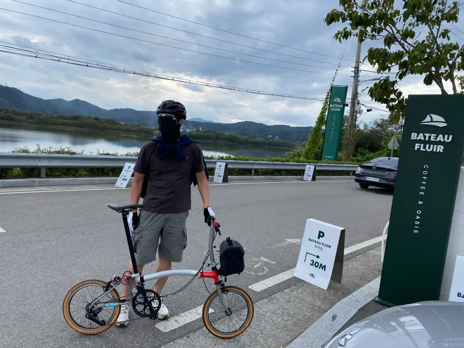 일상복 차림으로 브롬톤을 끌고 강변 카페 앞에 서 있는 라이더 — 브롬톤 장점인 복장 자유를 보여주는 실제 라이딩 사진