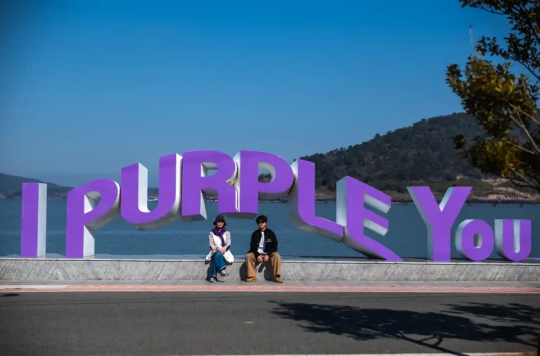 안좌도 퍼플섬 'I PURPLE YOU' 대형 조형물 앞 화강암 받침대에 두 사람이 나란히 앉아 있고, 뒤로 잔잔한 바다와 낮은 산 능선이 펼쳐지는 맑은 날 풍경