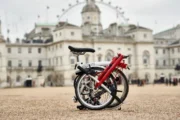 런던의 아이코닉한 건물을 배경으로 접힌 빨간색과 흰색 브롬톤 자전거가 지면에 세워져 있다.
